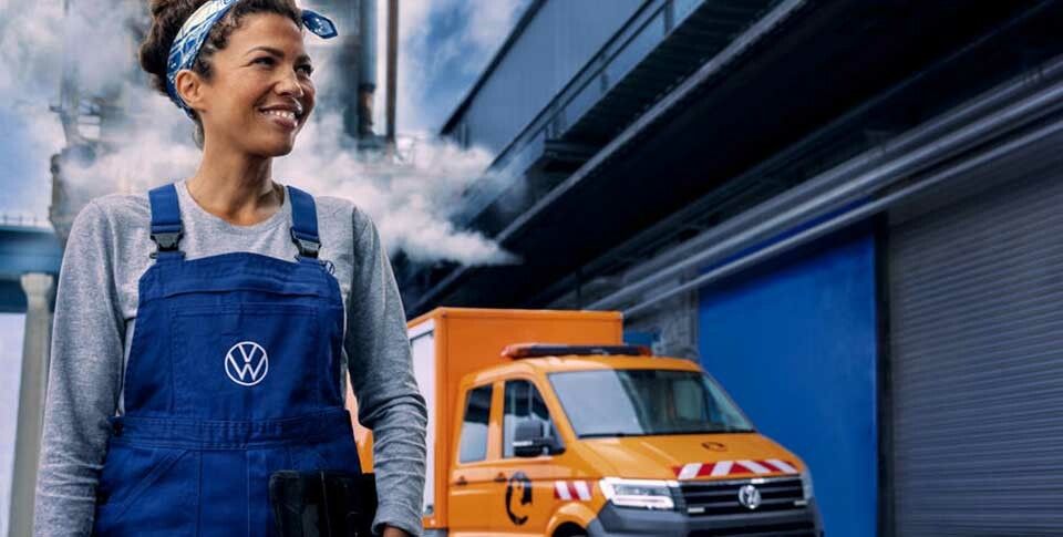 eine Volkswagen-Mitarbeiterin in blauer Arbeitslatzhose und orangem Nutzfahrzeug mit Warnmarkierung